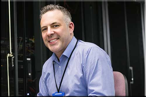 A picture of a sitting man wearing a button-down shirt with a conference pass smiling for the camera